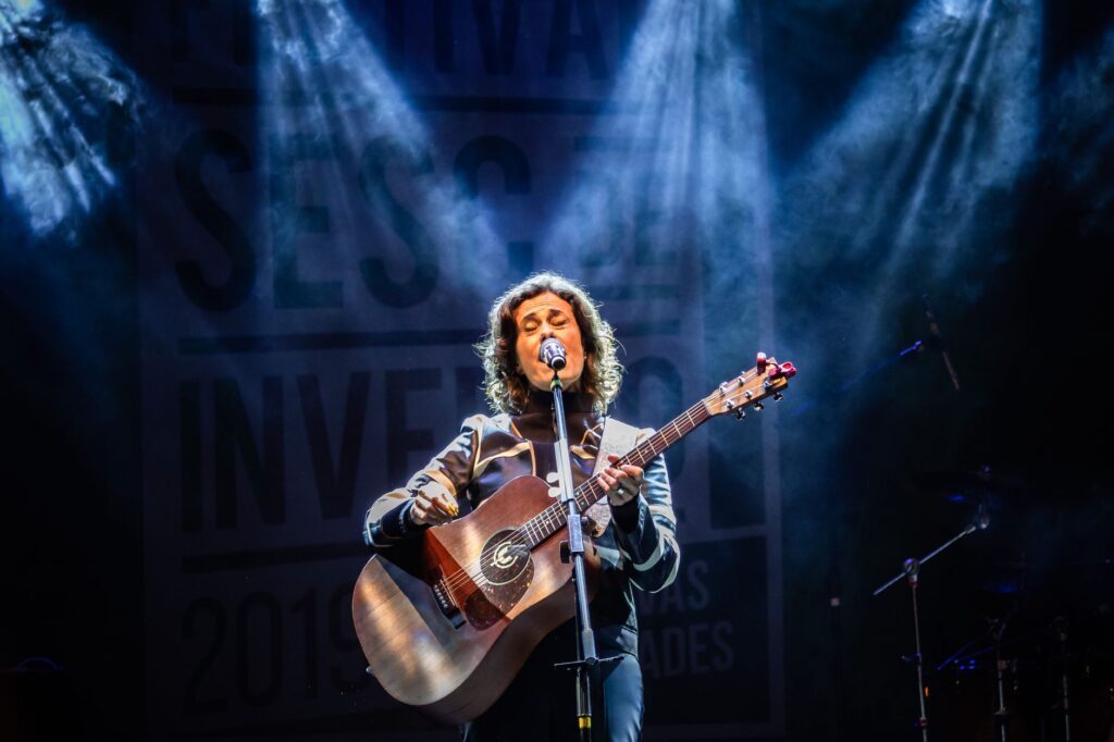 Musician performing live with guitar at Teresópolis festival, stage lit with colorful lights.