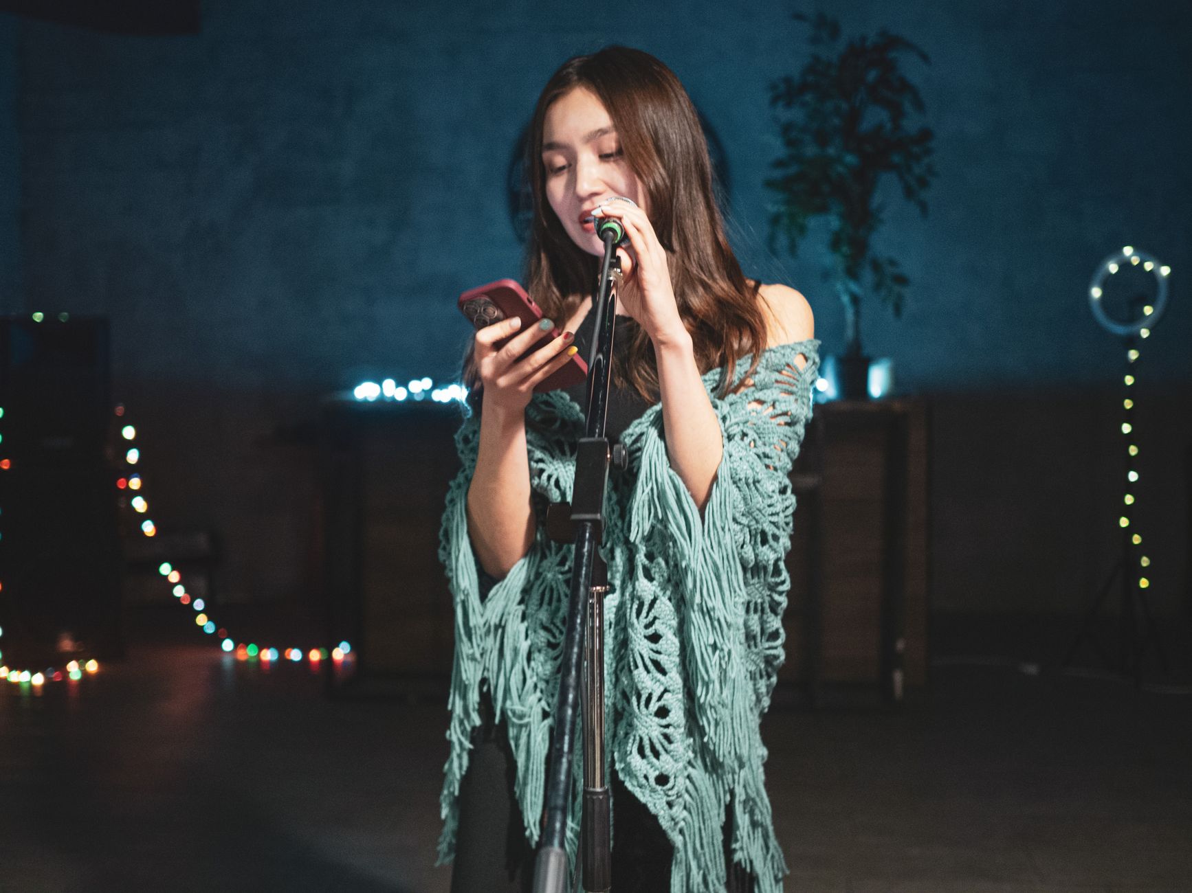 Young woman performs on stage, singing into a microphone, illuminated by stage lights and string lights.