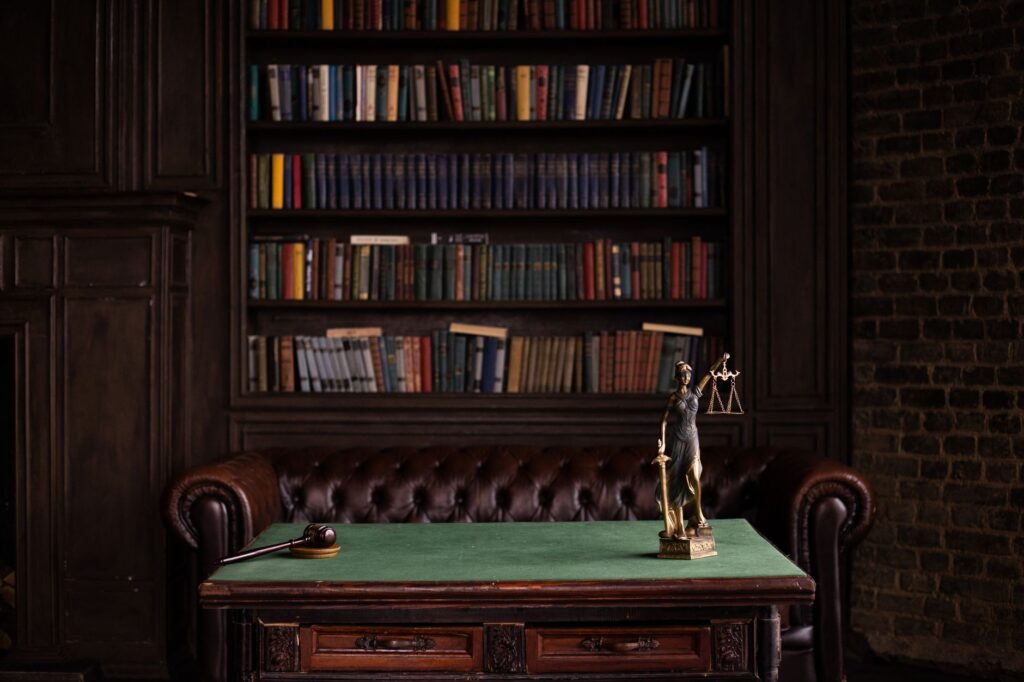 A classic study room with shelves of books, a gavel, and Lady Justice figurine on a green table.