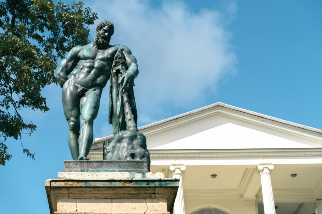 Bronze statue of Hercules beside a neoclassical building under a blue sky.