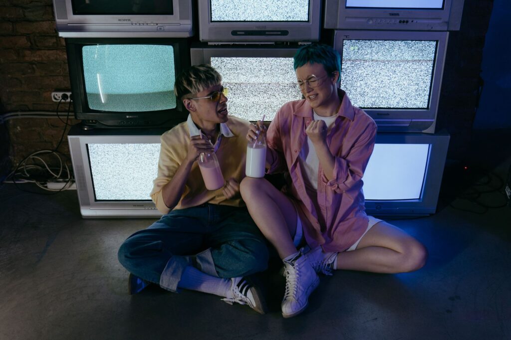 Two stylish teenagers sit with milkshakes amid vintage CRT TVs emitting static, exuding a retro and trendy vibe.
