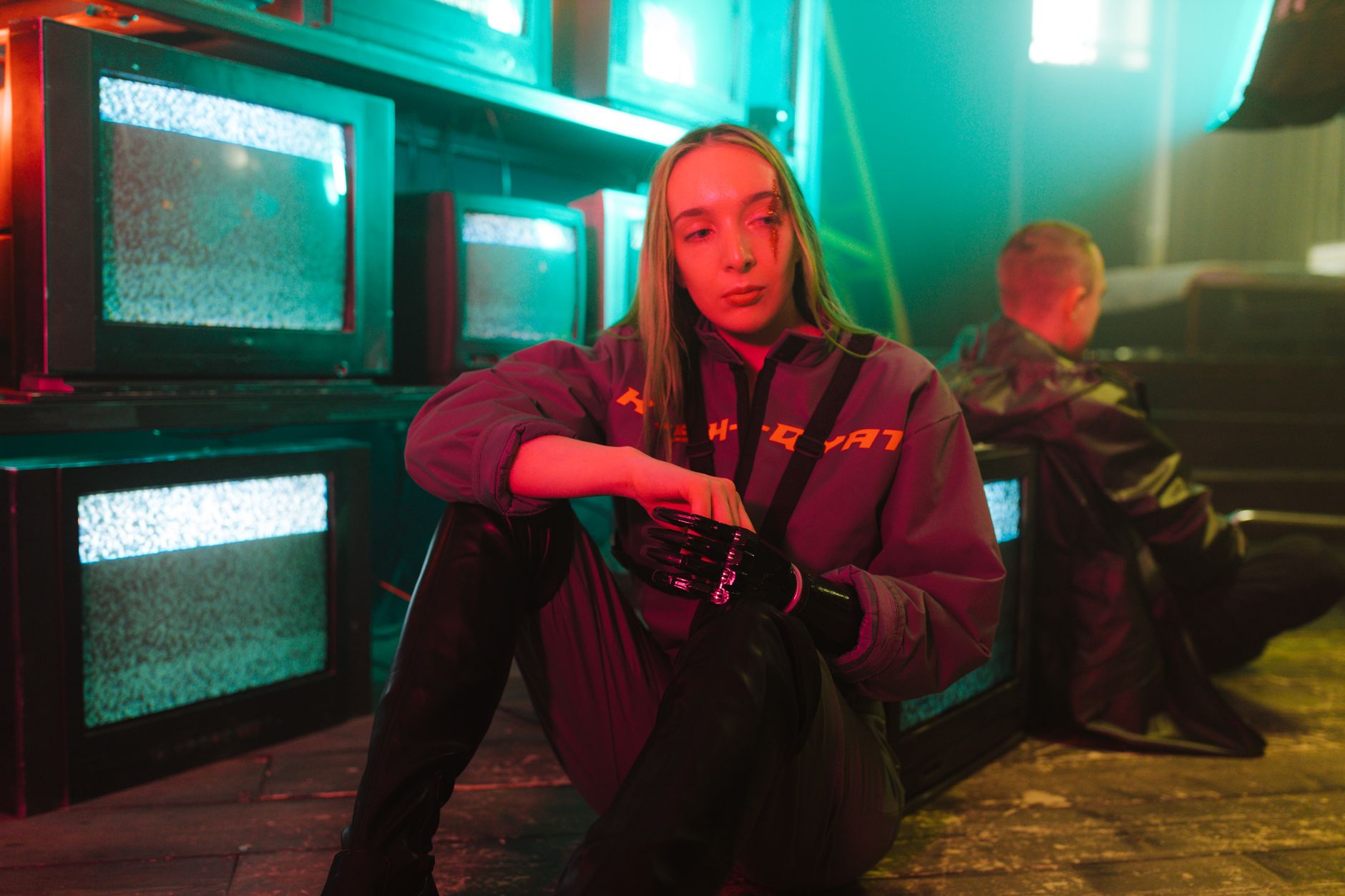 A woman with a prosthetic hand sits near old TVs in a cyberpunk-themed room.