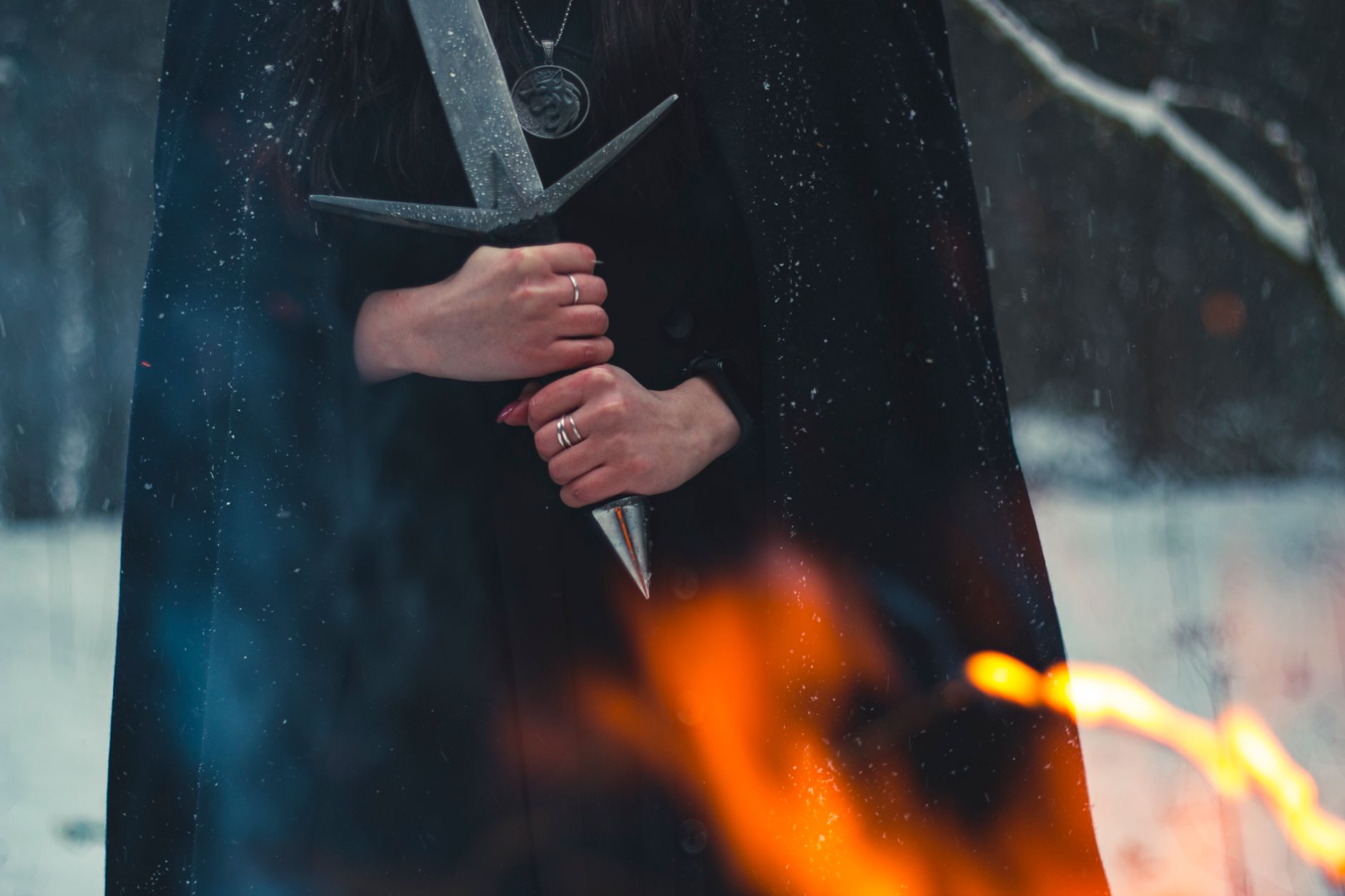 A cloaked figure holds a sword amidst snow and flames, evoking mystery.