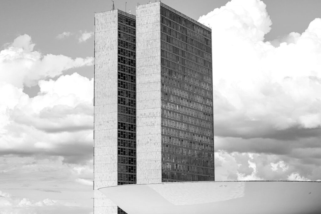 Black and white of tall triangular old multi storey construction next to round roof of modern building under sky covered with clouds